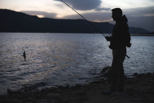 Man fishing on mountain lake. Sunrise low light. Casual clothes