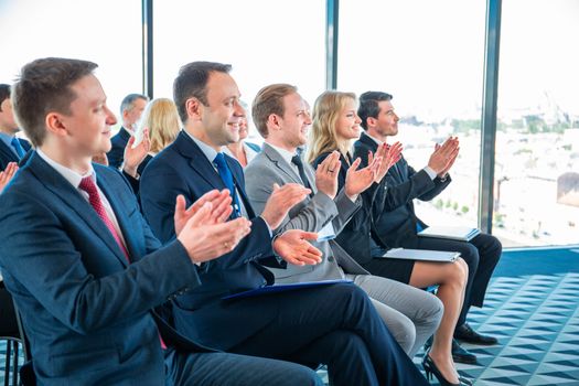 Group of happy business people in formalwear sitting at the chairs in conference hall and applauding to speaker