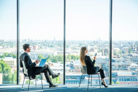 Business man with laptop and business woman with smartphone sitting by the window in panoramic office