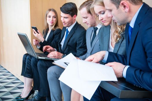 Group of business people is sitting in a row in an office lobby and waiting for the job interview or working with documents and laptops