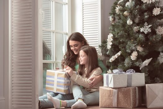 Happy mother and her little daughter unpack gift box near the Christmas tree at home