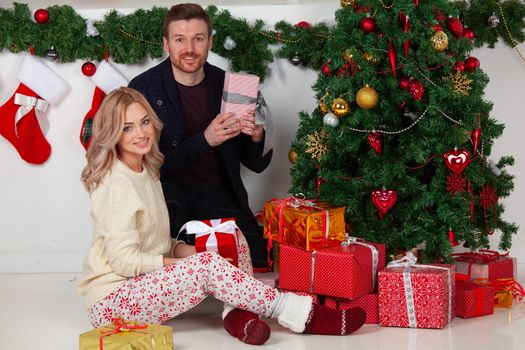 Couple in love sitting next to a nicely decorated Christmas tree, holding Christmas gifts and smiling