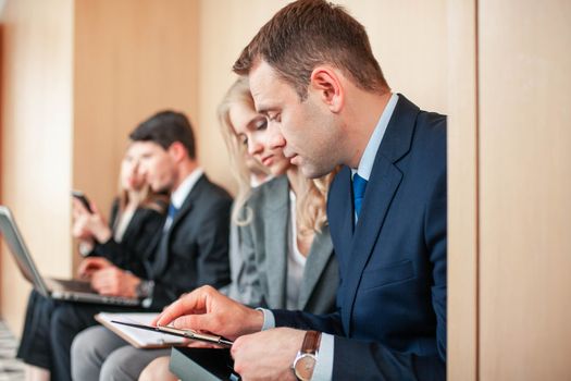Group of business people is sitting in a row in an office lobby and waiting for the job interview or working with documents and laptops