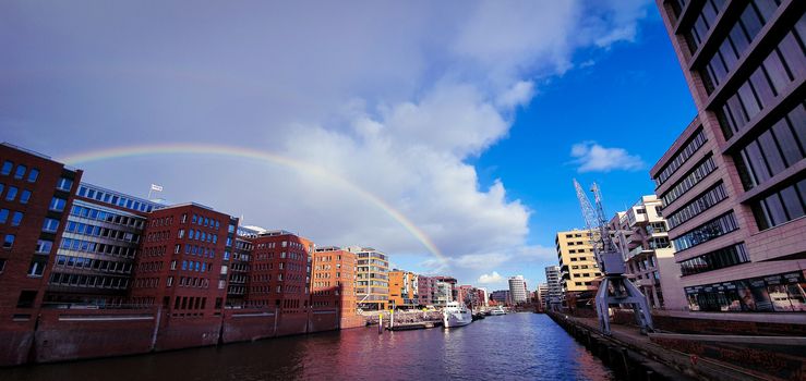 Beautiful rainbow over Hamburg canal. Beautiful wide angle view from the Kaiserkai bridge on the water canal at the Speicherstadt warehouse district near the Elbphilharmony concert hall. Buildings constructed with steel and brick.