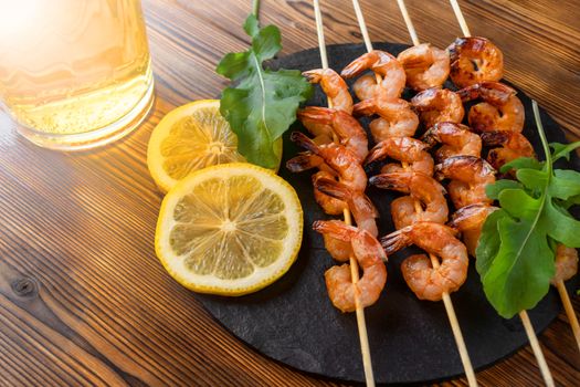 Shrimp fried on skewers with arugula, lemon on a black slate dish and beer in a glass on a wooden table.
