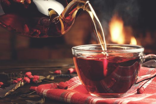 Pouring hot tea with hawthorn into a glass cup in a room with a burning fireplace in the background.