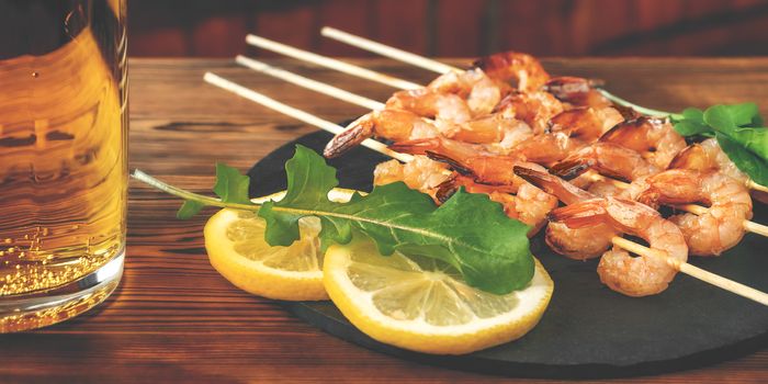 Shrimp fried on skewers with arugula, lemon on a black slate dish and beer in a glass on a wooden table.