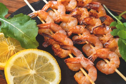 Shrimp fried on skewers with arugula and lemon on a black slate dish on a wooden table, top view, flat lay, close up.