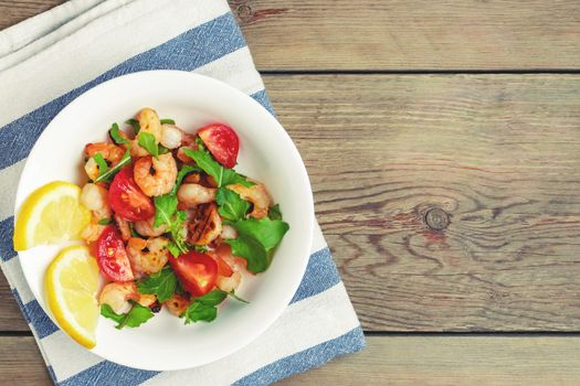 Fresh homemade salad of shrimp, arugula and tomato in a white plate on a wooden table, top view, flat lay, copyspace.