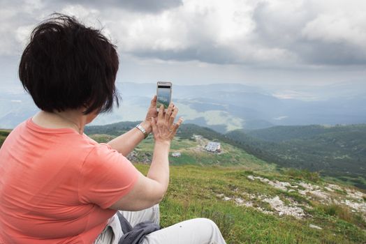 Senior woman taking photos with smartphone on the mountain. Dramatic sky