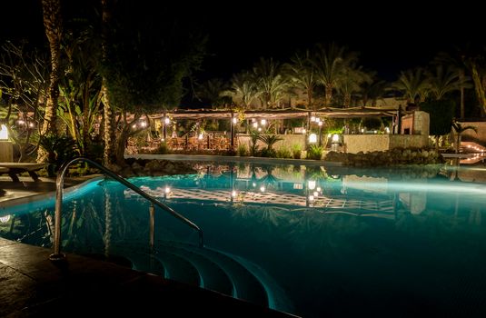 Sharm El Sheikh, Egypt - 02 06 2018: night in the hotel aquamarine pool empty armchairs in the bar palm trees light lanterns reflected in the water