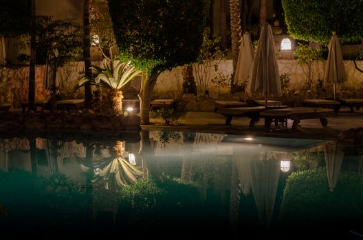 Sharm El Sheikh, Egypt - 02 06 2018: night in the hotel aquamarine pool folded umbrellas empty wooden deck chairs palm trees and decorative trees lantern light reflected in the water