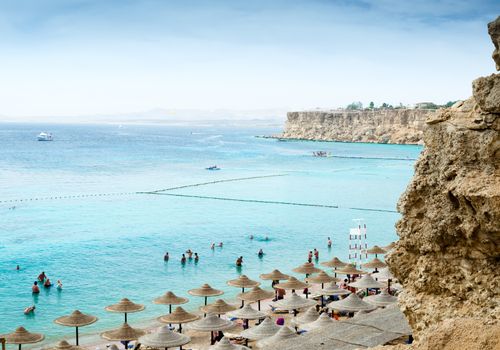 view from the top of the beach with umbrellas of reeds boats and people standing in blue water on a partly cloudy day