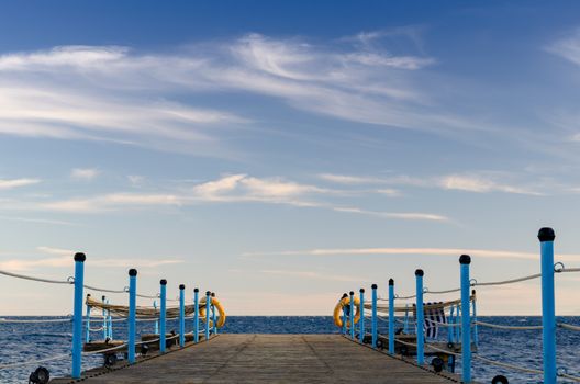 wooden platform with blue posts with ropes and orange lifebuoys on the background of the sea and sky with clouds