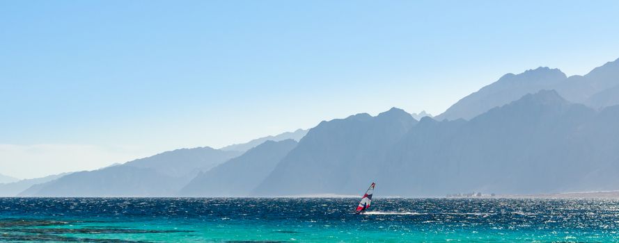 surfer rides in the Red Sea against the backdrop of high rocky mountains and a blue sky with clouds in Egypt Dahab