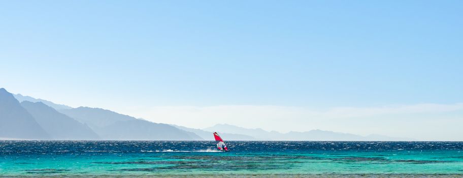 surfer rides in the Red Sea against the backdrop of high rocky mountains and a blue sky with clouds in Egypt Dahab