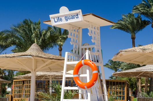 wooden lifeguard tower with orange lifebuoy on the beach against the backdrop of palm trees and reed parasols