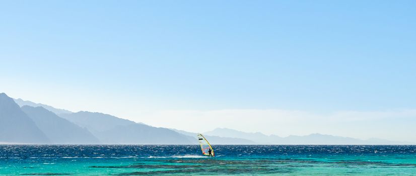 surfer rides in the Red Sea against the backdrop of high rocky mountains and a blue sky with clouds in Egypt Dahab