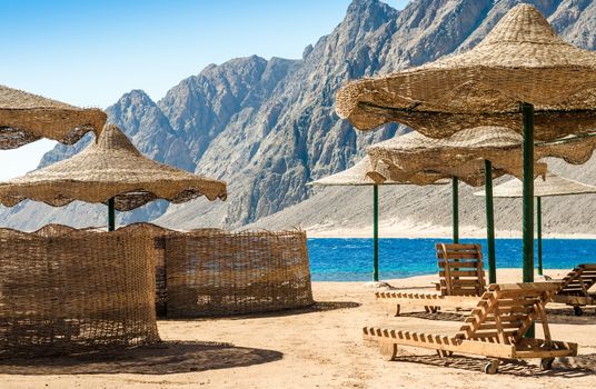 beach umbrellas and wooden lounge chairs on the sand of the beach against the backdrop of the sea and high rocky mountains