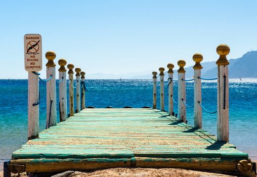 wooden pier with columns and a warning sign against a blue sea and mountains on the horizon