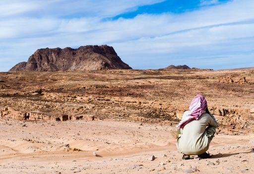 Bedouin in white red scarf in the desert against the backdrop of mountains and blue sky