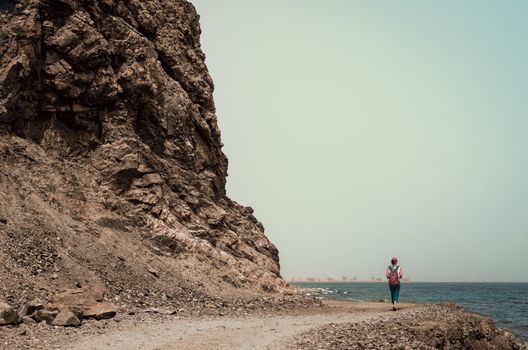 young girl tourist in a red cap with a backpack goes along the rocky shore of the Red Sea in Egypt