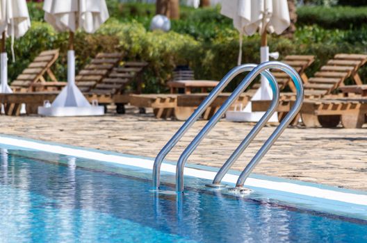 handrails by the pool with blue water on the background of sun loungers and folded white umbrellas