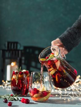 Female hand pours winter sangria in glasses with fruit slice, cranberry and rosemary. Dark christmas holiday background with candle, decoration lighting chain. Copy space for text or design. Vertical