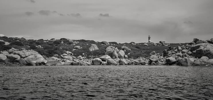 Lighthouse nestled in a natural promontory developed in horizontal liea under a dark and cloudy sky taken from in the middle of the sea. Image in black and white.
