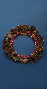 Christmas wreath of pine cones and glowing lights garland on blue background top view flat lay copy space text