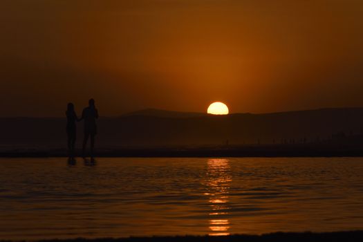 A young couple watches from the edge of a tide pool as the sun sets over the horizon, Mossel Bay, South Africa