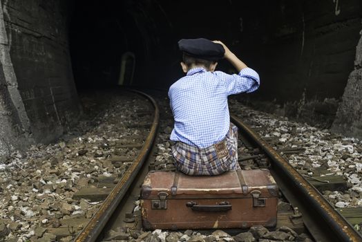Child in vintage clothes sits on railway road in front of a tunnel.