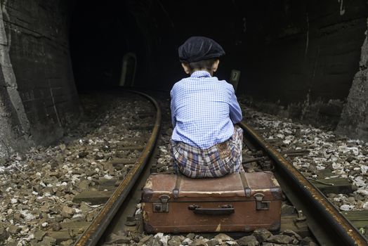 Child in vintage clothes sits on railway road in front of a tunnel.