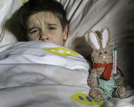 Sick child in bed with teddy bear. Measuring the temperature with a thermometer.