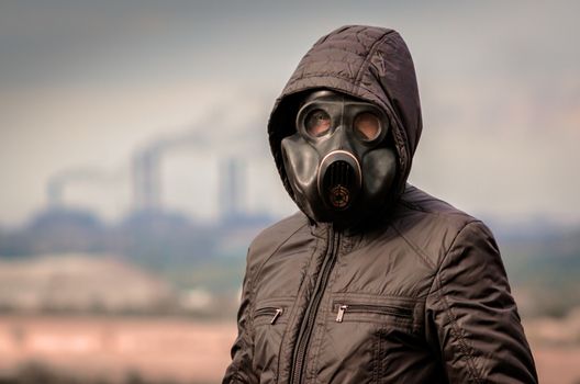 portrait of a man in a gas mask and a hood against the background of smoke from factory pipes close up