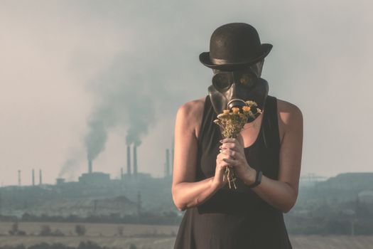 young girl in a black dress and a gas mask with flowers on the background of smoking factory chimneys close up