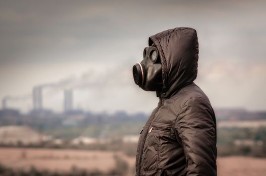 man in a gas mask and a hood amid smoke from factory pipes close up