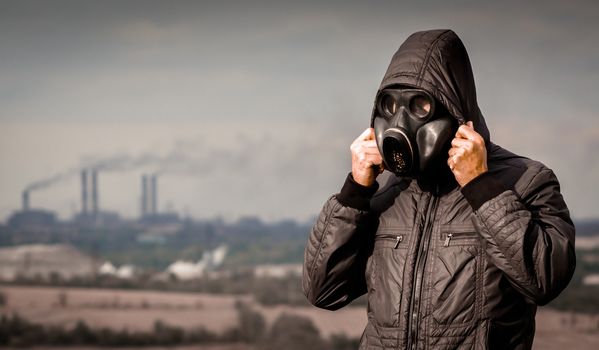 man in a gas mask and a hood against the background of smoke from factory pipes close up