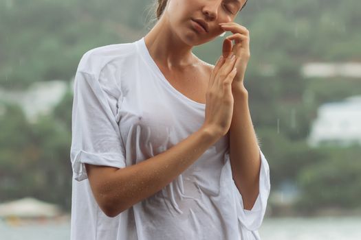 Attractive young female in while blouse and panties embracing herself and keeping eyes closed while standing near water on windy day