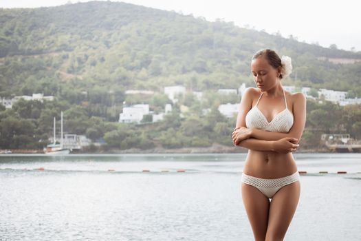 Sensual young lady in knitted swimwear embracing herself and keeping eyes closed while standing near amazing calm sea