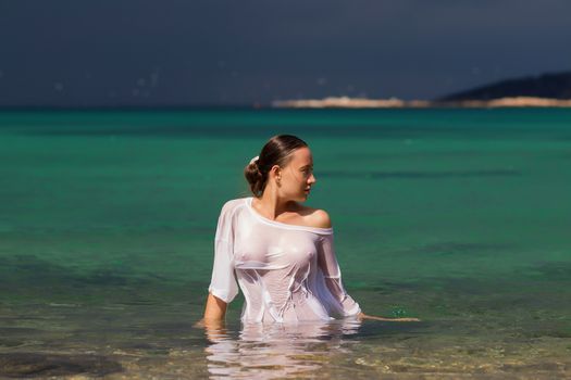 Sensual young female in wet blouse standing in clean sea water and looking away