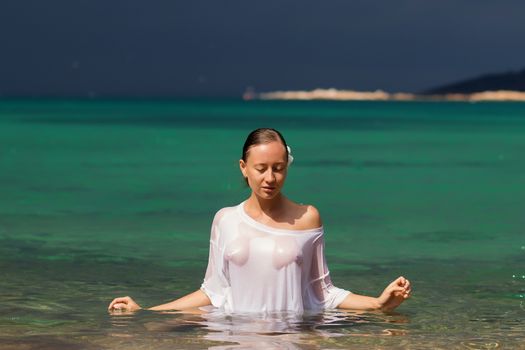 Sensual young female in wet blouse standing in clean sea water and looking away