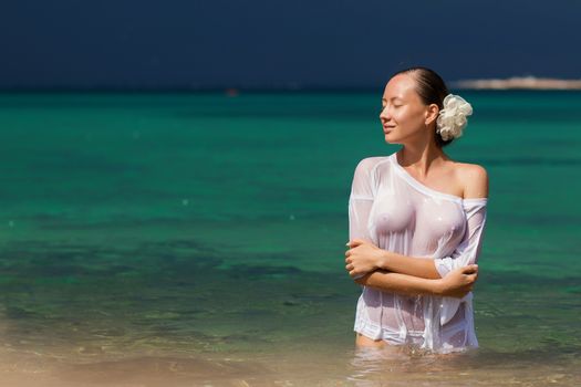 Sensual young female in wet blouse standing in clean sea water and looking away