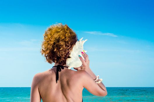 young woman with red hair with a big sea shell and colorful manicure on the background of the sea and blue sky
