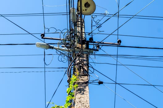 pole with electric wires and street lighting with green leaves against a blue sky background