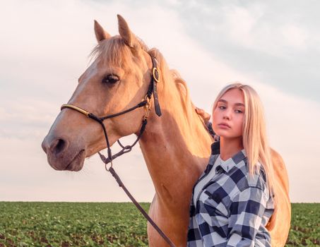 young blonde girl with a beige horse on the ranch on the background of a green field and evening sky