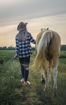 young blonde girl in a hat and a plaid shirt walks with a horse in the evening on a farm in the village