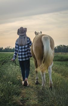 young blonde girl in a hat and a plaid shirt walks with a horse in the evening on a farm in the village