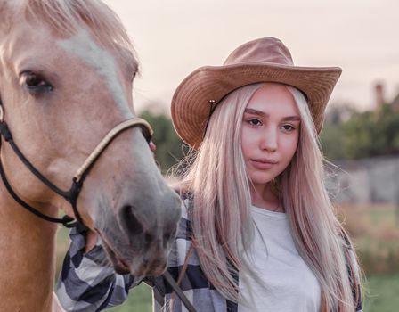 portrait of a young blonde girl dressed in a cowboy hat with a beige horse on a ranch closeup