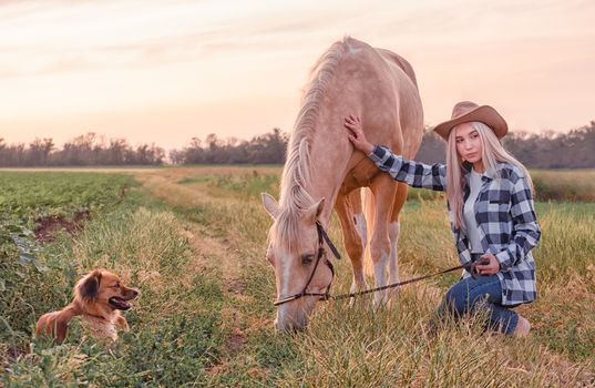 young blonde girl dressed in a cowboy hat and blue jeans with a horse and dog on the ranch